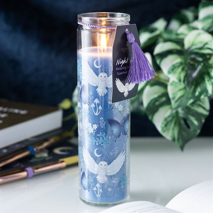 Candle in a glass jar with decorative label and tassel, placed on a surface with books and plants in the background.