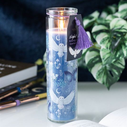 Candle in a glass jar with decorative label and tassel, placed on a surface with books and plants in the background.