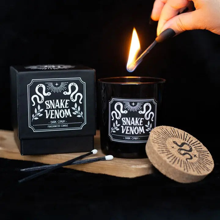 Candle labeled 'Snake Venom' being lit with a match, next to its packaging and lid.